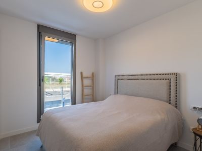 Chambre lumineuse avec lit, tête de lit en tissu clouté et armoire intégrée. Grande fenêtre offrant un accès extérieur et un sol carrelé gris.
