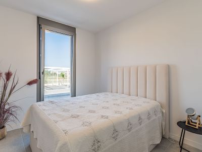 Chambre lumineuse avec un lit et une tête de lit rembourrée beige. Grande fenêtre du sol au plafond offrant une vue extérieure. Sol carrelé gris et plante décorative.