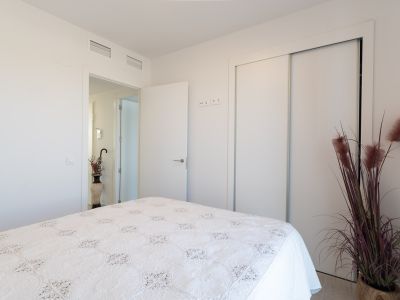 Chambre lumineuse avec un lit orné d'un couvre-lit blanc brodé. Elle dispose d'une armoire encastrée à portes coulissantes et de bouches de climatisation au plafond. Une plante décorative agrémente l'espace.