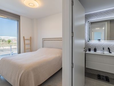 Chambre principale avec lit à tête de lit rembourrée et accès direct à une terrasse extérieure. Une salle de bain attenante avec meuble-lavabo suspendu et miroir rétroéclairé est visible.