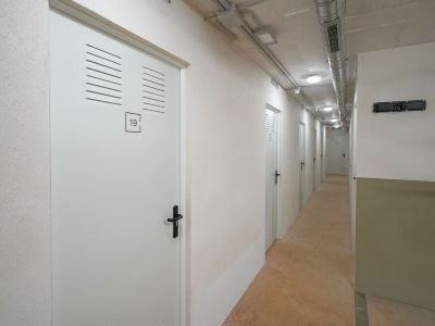 Couloir de stockage avec des portes blanches à persiennes, numérotées, et poignées noires. Le plafond révèle des conduits apparents et un éclairage encastré, au-dessus d'un sol en béton.