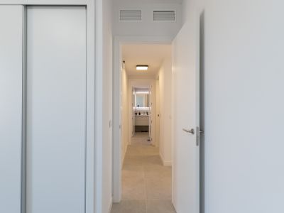 Couloir lumineux avec placard coulissant blanc et porte ouverte. Le carrelage clair mène à une salle de bain moderne avec miroir rétroéclairé et meuble-lavabo contemporain. Grilles de ventilation au plafond.