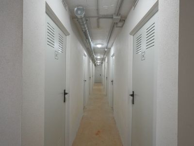 Couloir utilitaire vers des unités de stockage individuelles, avec portes blanches numérotées et fentes d'aération. Plafond en béton apparent avec conduits industriels et éclairage encastré, murs texturés.
