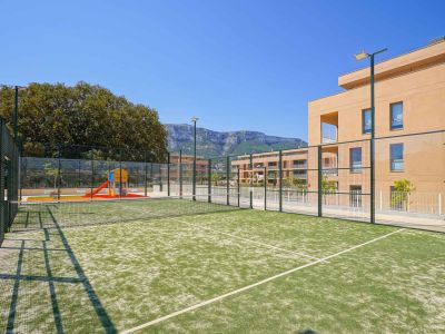 Court de padel en gazon synthétique vert, clôturé et éclairé, jouxtant une aire de jeux pour enfants et des bâtiments résidentiels modernes avec vue sur les montagnes.