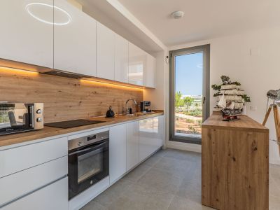 Cuisine moderne avec armoires blanches laquées, plan de travail et crédence effet bois. Équipée d'appareils intégrés, d'un éclairage LED et d'une fenêtre donnant sur l'extérieur paysagé.