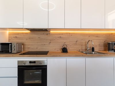 Cuisine moderne avec des armoires blanches laquées, un plan de travail et une crédence effet bois, et un éclairage LED sous les meubles hauts. Four encastré, plaque à induction et micro-ondes.