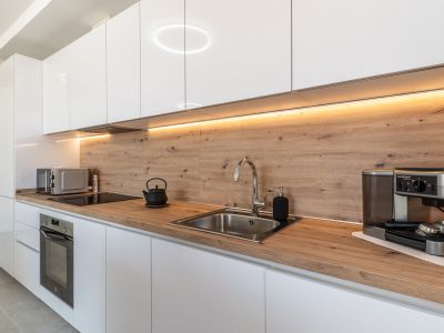 Cuisine moderne avec meubles blancs laqués, plan de travail et crédence aspect bois clair. Elle intègre un four, une plaque à induction et un évier en inox, soulignée par un éclairage LED sous les placards.