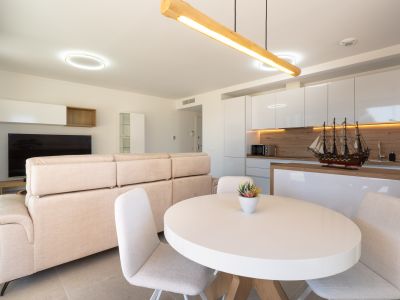 Espace salon-salle à manger ouvert avec cuisine intégrée. Il présente un canapé clair, une table ovale blanche et une cuisine moderne avec des meubles laqués blancs et des accents boisés.