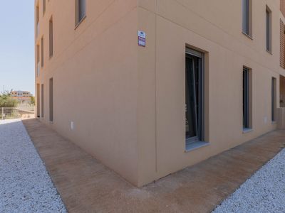 Extérieur d'un bâtiment moderne avec façade en stuc beige clair et fenêtres à cadres sombres. Le sol combine un chemin en béton texturé et un aménagement paysager de gravier blanc, avec alarme visible.