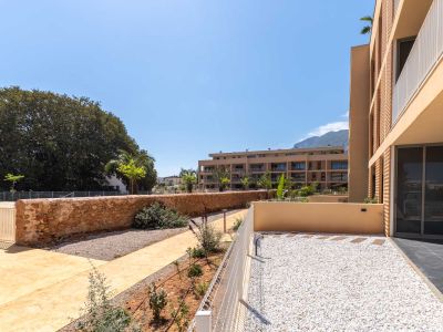 Jardin privé en rez-de-chaussée avec gravier blanc et dalles, délimité par une clôture basse et un mur en pierre. Vues sur les bâtiments modernes et les montagnes lointaines.