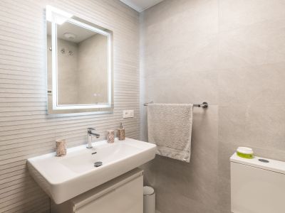 Meuble-lavabo de salle de bain moderne avec lavabo rectangulaire blanc, robinet mural, miroir éclairé et carrelage mural gris texturé.