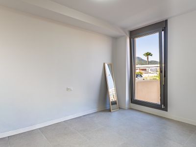 Pièce vide avec murs blancs et carrelage gris, prête à être aménagée. Grande fenêtre offrant une lumière naturelle généreuse et des vues sur un palmier et les montagnes.