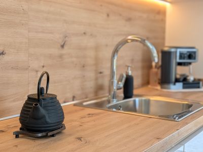 Plan de travail et crédence de cuisine modernes en imitation bois clair. Évier en acier inoxydable avec robinet chromé, éclairage LED sous les meubles et théière en fonte au premier plan.