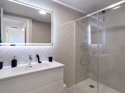 Salle de bain contemporaine avec meuble-lavabo blanc, miroir éclairé et carrelage mural à motif chevrons. La douche vitrée est équipée d'une pomme de douche pluie et de robinetterie chromée.