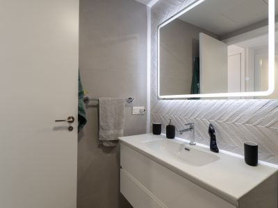 Salle de bain moderne avec meuble-lavabo suspendu blanc, robinetterie chromée et miroir lumineux. Caractérisée par des carreaux muraux texturés à motif chevrons.