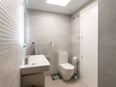 Salle de bain moderne carrelée en gris clair avec lavabo suspendu et toilettes. Un miroir éclairé et un mur texturé apportent du caractère.