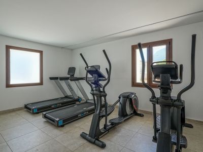 Salle de sport résidentielle équipée de deux tapis de course BH RCM Pro et deux vélos elliptiques. Elle dispose d'un sol carrelé, de murs blancs et de fenêtres à cadres en bois, l'une avec vue sur la montagne.
