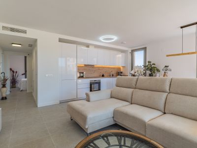 Salon-cuisine ouvert avec canapé sectionnel beige, cuisine moderne aux armoires blanches brillantes et crédence effet bois clair. Carrelage au sol et lumière naturelle.