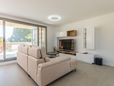 Salon lumineux avec un canapé sectionnel beige, carrelage gris clair et de grandes baies vitrées coulissantes donnant sur l'extérieur. Meuble TV contemporain avec rangements blancs et bois.