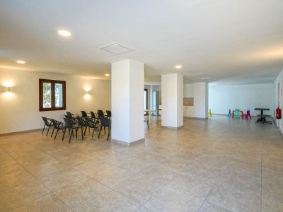 Vaste centre social commun avec carrelage clair, piliers porteurs et éclairage encastré. Comprend un espace de réunion avec chaises et un coin jeux coloré pour enfants.