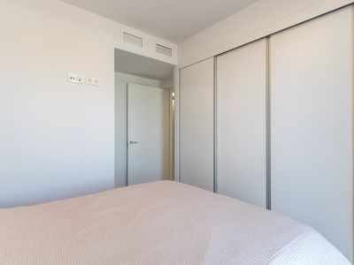 Vue de chambre à coucher avec une grande armoire intégrée à portes coulissantes blanches. Le mur dispose de prises électriques modernes et de grilles de ventilation au-dessus de la porte adjacente.