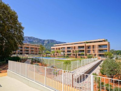 Vue extérieure du complexe résidentiel moderne avec deux bâtiments aux façades claires, une piscine commune et des jardins aménagés. Les montagnes sont visibles en arrière-plan.