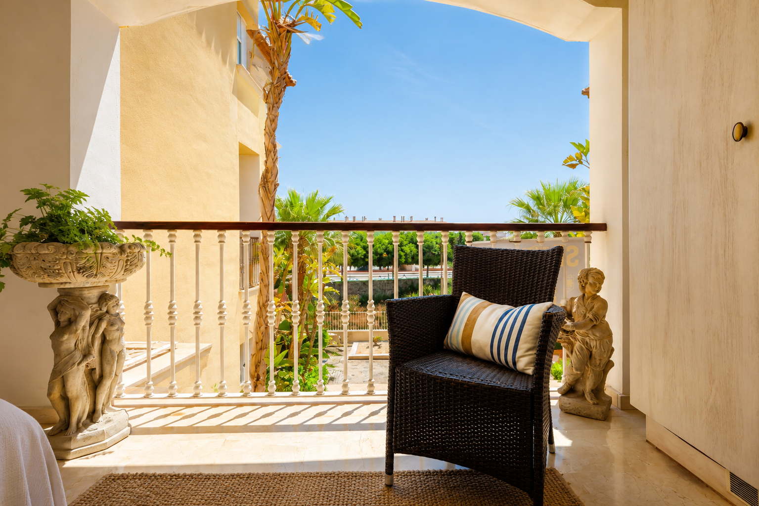 Agréable terrasse privée avec un fauteuil en osier foncé, sol en marbre clair et une balustrade ornée. Agrémentée de jardinières en pierre, elle offre des vues sur les palmiers et un ciel azur.