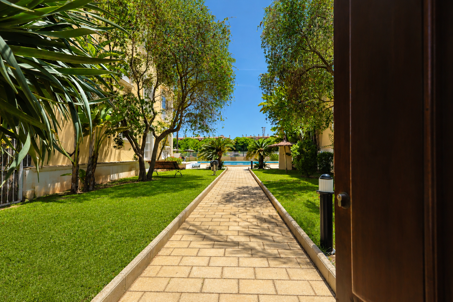 Chemin pavé traversant les jardins communs luxuriants, menant à la piscine et aux palmiers sous un ciel bleu. Vue depuis une porte ouverte.
