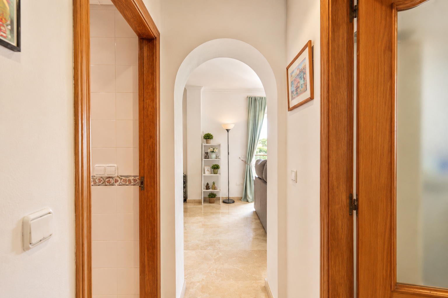 Couloir arqué avec sol en marbre clair, menant à un salon avec fenêtre et rideaux bleus doux. Des encadrements de porte en bois flanquent le passage, l'un révélant un mur carrelé.