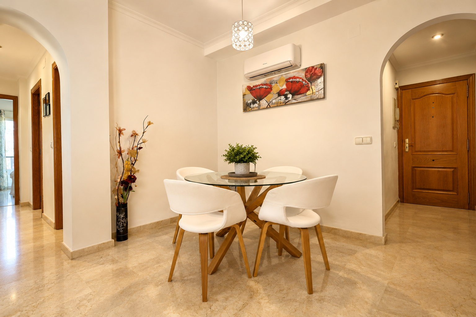 Espace repas compact avec une table ronde en verre à piètement bois, quatre chaises blanches et un sol en marbre clair. Un climatiseur mural et une œuvre florale sont visibles.