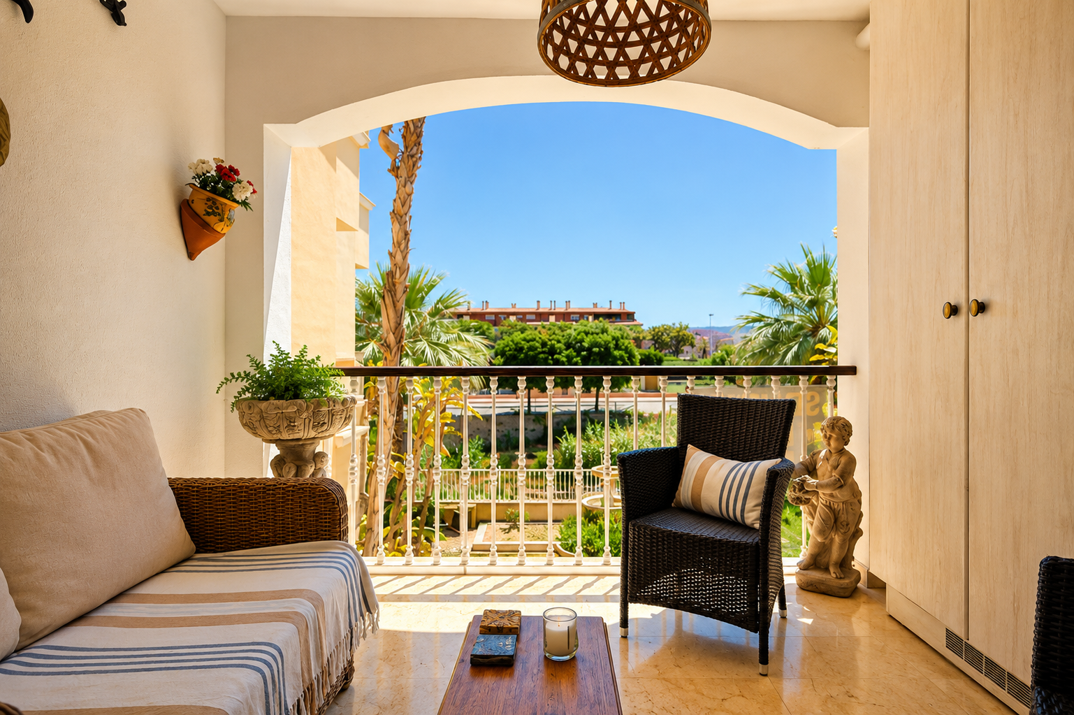 La terrasse privée, avec son sol effet marbre clair, offre un canapé et un fauteuil en osier. Une arche encadre des vues sur les palmiers, jardins et bâtiments lointains sous un ciel bleu, avec rangements intégrés.