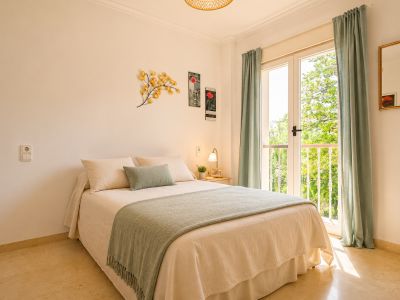 Chambre lumineuse avec lit double, sol en marbre clair et accès direct à un balcon offrant une vue verdoyante. Rideaux vert menthe et décoration murale.