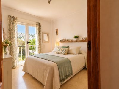 Chambre principale lumineuse avec accès direct au balcon, offrant des vues sur la ville et la mer. Caractérisée par son sol en marbre clair et son étagère en bois rustique au-dessus du lit.