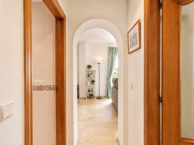 Couloir arqué avec sol en marbre clair, menant à un salon avec fenêtre et rideaux bleus doux. Des encadrements de porte en bois flanquent le passage, l'un révélant un mur carrelé.