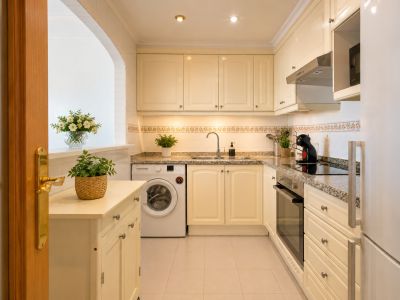 Cuisine fonctionnelle avec armoires crème, plans de travail en granit et électroménager intégré. Une ouverture arquée et une porte en bois encadrent l'espace.