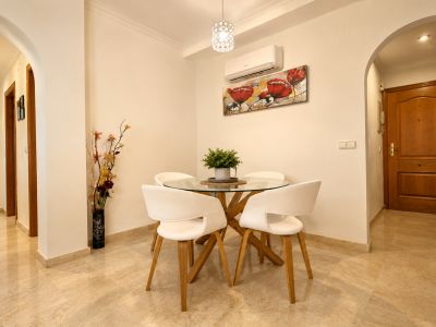 Espace repas compact avec une table ronde en verre à piètement bois, quatre chaises blanches et un sol en marbre clair. Un climatiseur mural et une œuvre florale sont visibles.