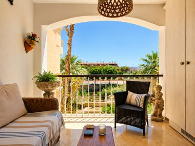 La terrasse privée, avec son sol effet marbre clair, offre un canapé et un fauteuil en osier. Une arche encadre des vues sur les palmiers, jardins et bâtiments lointains sous un ciel bleu, avec rangements intégrés.