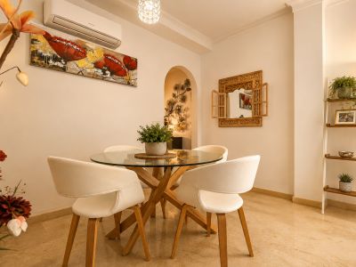 Salle à manger lumineuse avec table ronde en verre et piètement en bois, chaises blanches, sol en marbre clair et un miroir en bois sculpté, complétée par une étagère décorative.