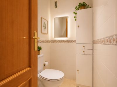 Salle de bain fonctionnelle avec murs carrelés crème et frise décorative, toilettes blanches et miroir rétroéclairé. Un meuble de rangement blanc à poignées dorées et une porte en bois sont visibles.