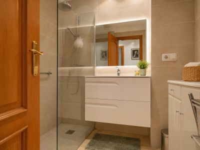 Salle de bain moderne avec meuble-lavabo suspendu blanc et miroir éclairé. Elle dispose d'une douche à l'italienne vitrée, de murs carrelés beiges et d'un sol en marbre clair.