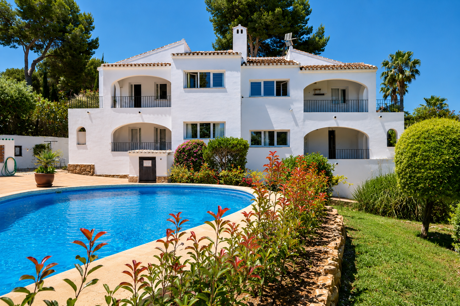 Bâtiment blanc de style méditerranéen avec plusieurs balcons arqués et balustrades noires. Une grande piscine courbe et des jardins luxuriants sont visibles sous un ciel bleu clair.