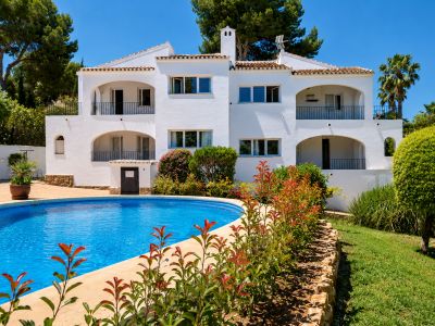 Bâtiment blanc de style méditerranéen avec plusieurs balcons arqués et balustrades noires. Une grande piscine courbe et des jardins luxuriants sont visibles sous un ciel bleu clair.
