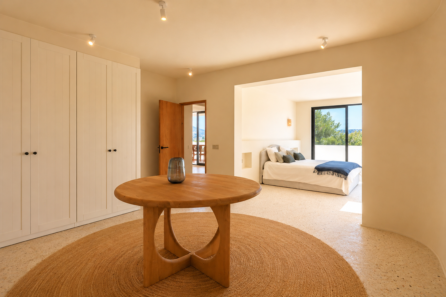 Cet espace de transition lumineux comprend une table ronde en bois sur un tapis en jute, flanquée d'armoires blanches intégrées. Il mène à une chambre avec vue sur la verdure.
