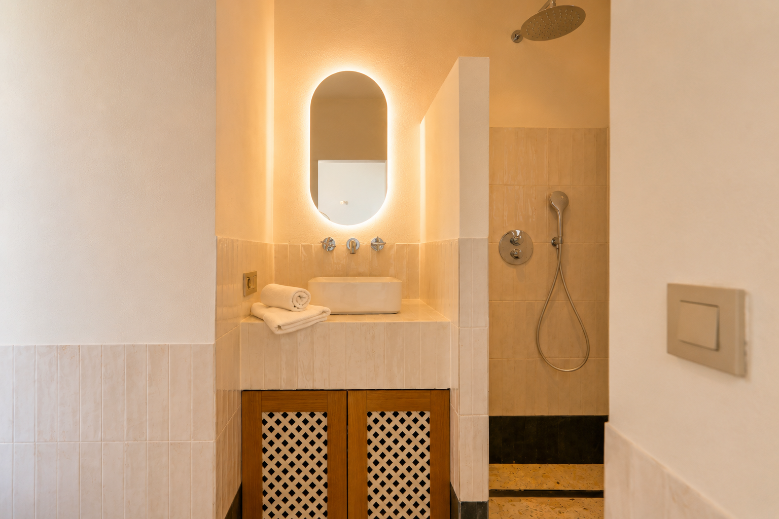 Cette salle de bain élégante présente une vasque, un miroir ovale rétroéclairé et un meuble en bois ajouré. La douche attenante est équipée d'une pomme de douche pluie.