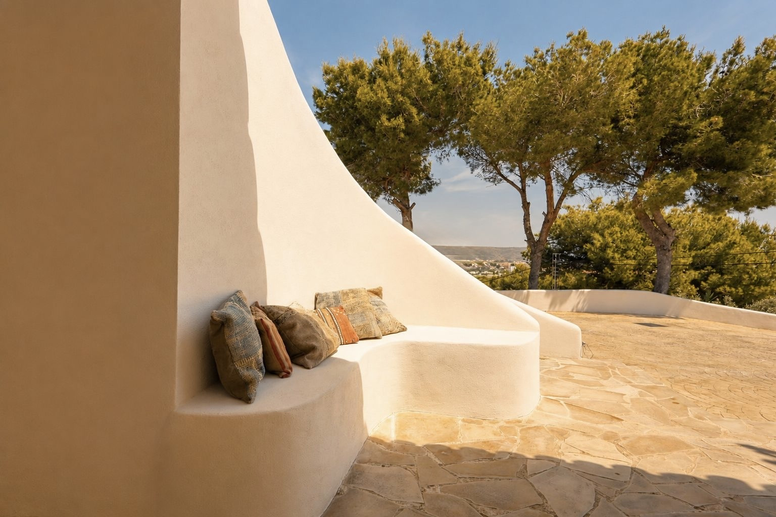 Cette terrasse extérieure offre un banc en stuc intégré aux courbes douces, agrémenté de coussins bohèmes, sur un patio en dalles irrégulières. Des pins et une vue lointaine sur le paysage sont visibles.