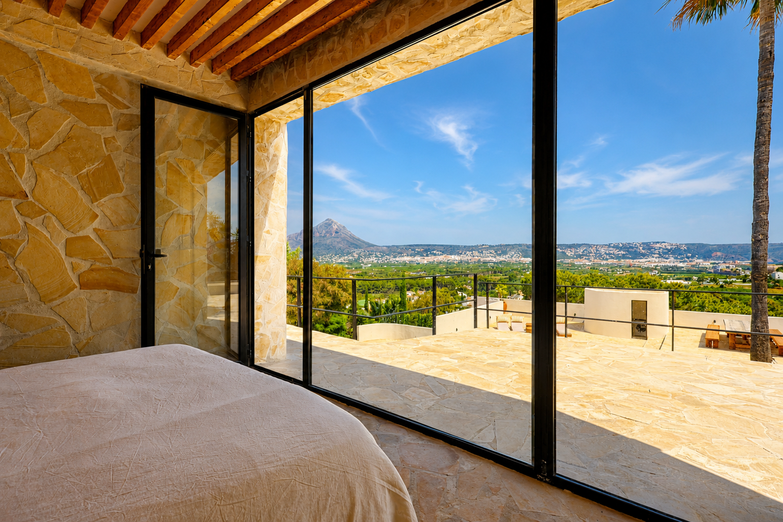 Chambre avec murs en pierre naturelle et poutres en bois, s'ouvrant sur une terrasse avec vues panoramiques sur le Montgó et la baie de Jávea.