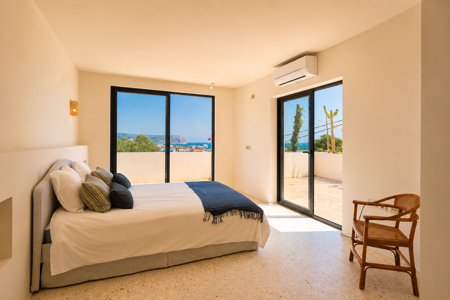 Chambre lumineuse avec sol en terrazzo clair et grandes portes coulissantes vitrées menant à une terrasse privée. Offre des vues étendues sur la mer et la côte lointaine.