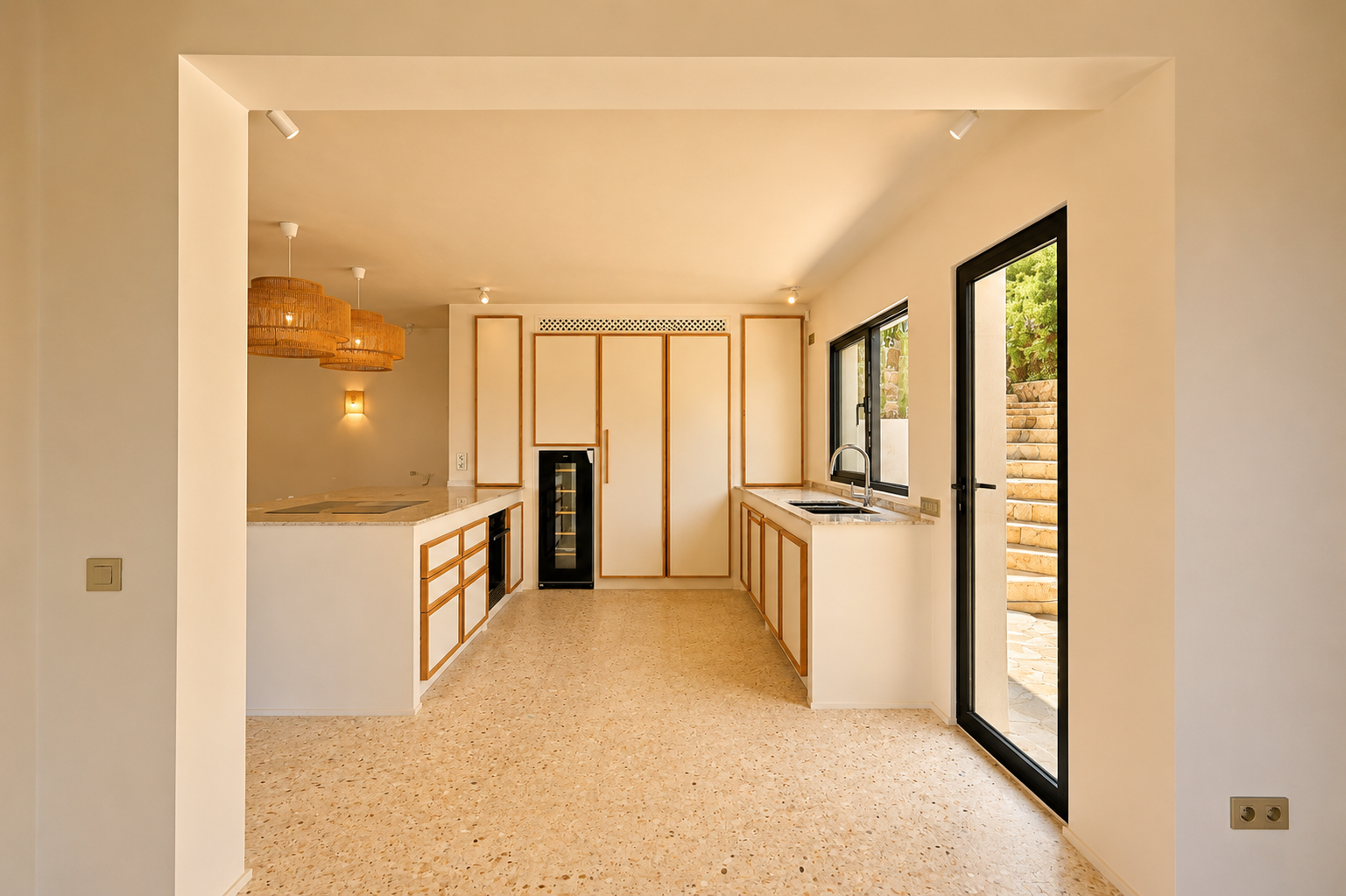Cuisine moderne avec des armoires bordées de bois clair, un sol terrazzo et un grand îlot avec table de cuisson intégrée. Des suspensions en rotin éclairent la pièce.