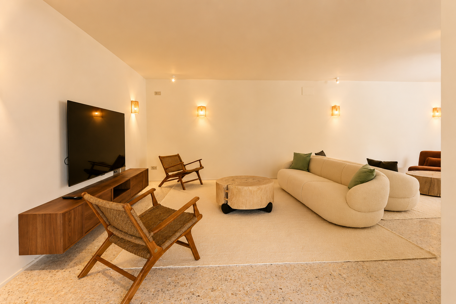 Espace de vie contemporain avec canapé incurvé crème, table basse rustique en bois et fauteuils en osier sur sol en terrazzo, éclairé par des appliques murales chaleureuses.