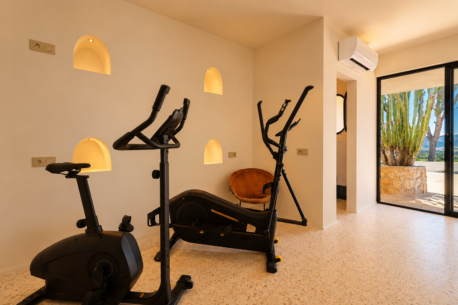 Espace fitness équipé d'un vélo d'appartement et d'un elliptique, avec des niches murales arquées illuminées et vue sur un jardin de cactus et les montagnes.